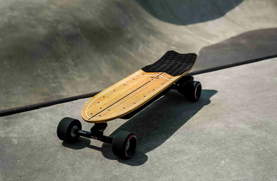 An electric skateboard with a wooden deck and black wheels is positioned on a concrete surface, casting a shadow.