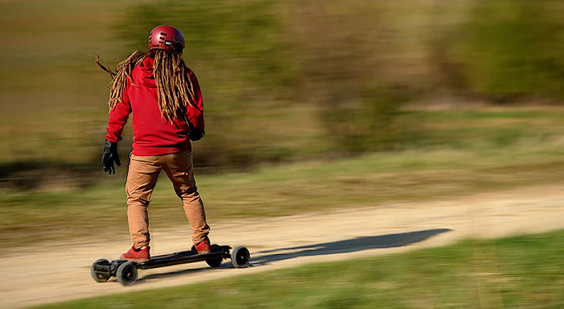 A person with dreadlocks, wearing a red helmet and red shoes, is riding an electric skateboard on a dirt path.