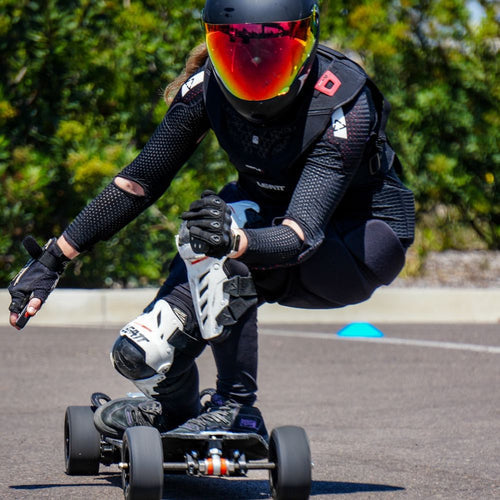 A person in full protective gear, including a helmet and knee pads, is riding a low-profile electric skateboard.